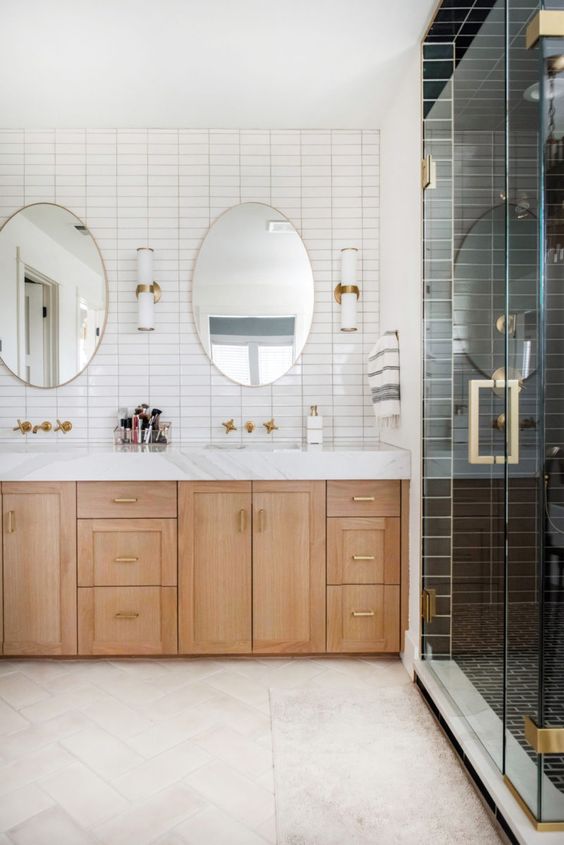 a two-tone bathroom with white and black stacked tiles, a light-stained vanity, oval mirrors, brass fixtures and sconces
