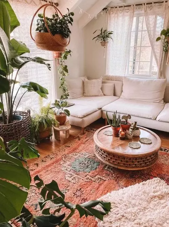 a very cozy boho sunroom with a white sectional, a wooden carved table, lots of potted greenery, layered rugs and macrame curtains