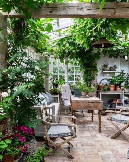 a vintage rustic sunroom with wooden furniture, printed textiles, potted greenery and blooms and lots of greenery