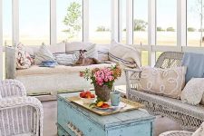 a vintage sunroom with all glazed walls, white wicker furniture, a blue shabby chic chest, printed and embroidered pillows and blooms in a vase