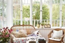 a vintage sunroom with white planked walls, wicker furniture, a bold rug, blankets and pillows, potted blooms
