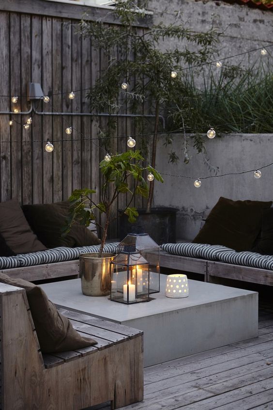 a welcoming Scandi backyard with wooden benches with printed upholstery and black pillows, a concrete coffee table and string lights