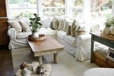 a welcoming farmhouse sunroom with a white L-shaped sofa, some vintage tables, baskets, neutral textiles and potted greenery