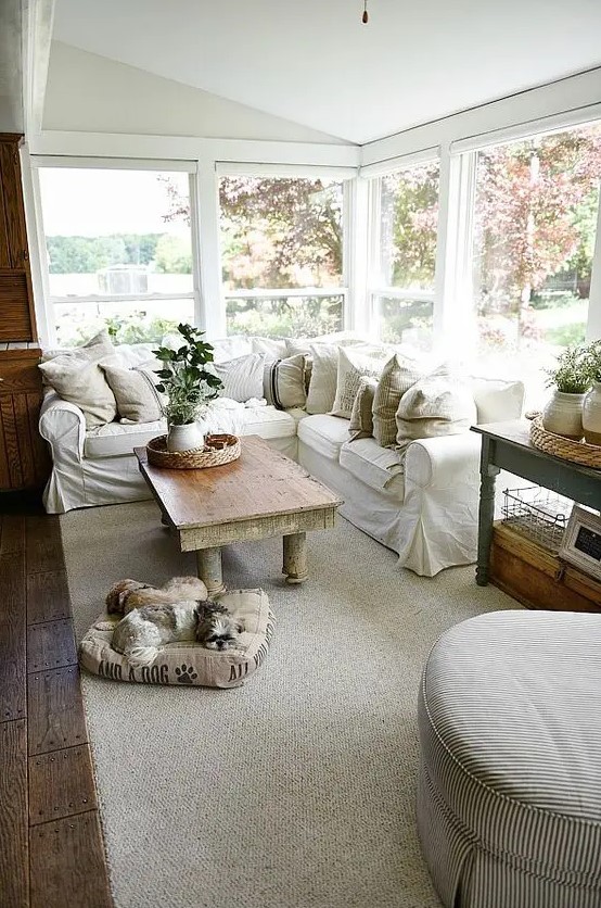 a welcoming farmhouse sunroom with a white L shaped sofa, some vintage tables, baskets, neutral textiles and potted greenery