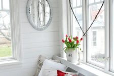 a white Scandi sunroom with planked walls, a built-in bench with printed pillows, crates for storage, tulips in a jug and a heart