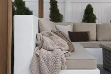 a white concrete corner sofa with neutral upholstery and neutral pillows and a wicker coffee table and potted plants