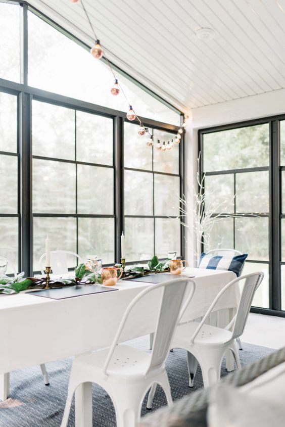 an airy and light-filled Scandinavian sunroom with a long table, white metal chairs, greenery and string lights is a gorgeous dining space