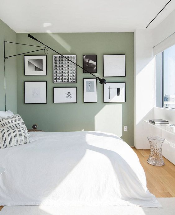 an airy minimalist bedroom with white and green walls, a white bed with neutral bedding, a windowsill desk and a neutral stool, a black and white gallery wall