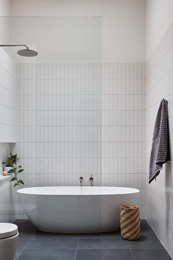 an airy neutral bathroom with stacked white tiles, an oval tub, a basket, a potted plant and a toilet, a glass shower space divider