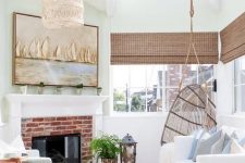 an elegant coastal sunroom with white and light blue furniture, woven shades, a brick fireplace, a woven lamp and a coastal artwork