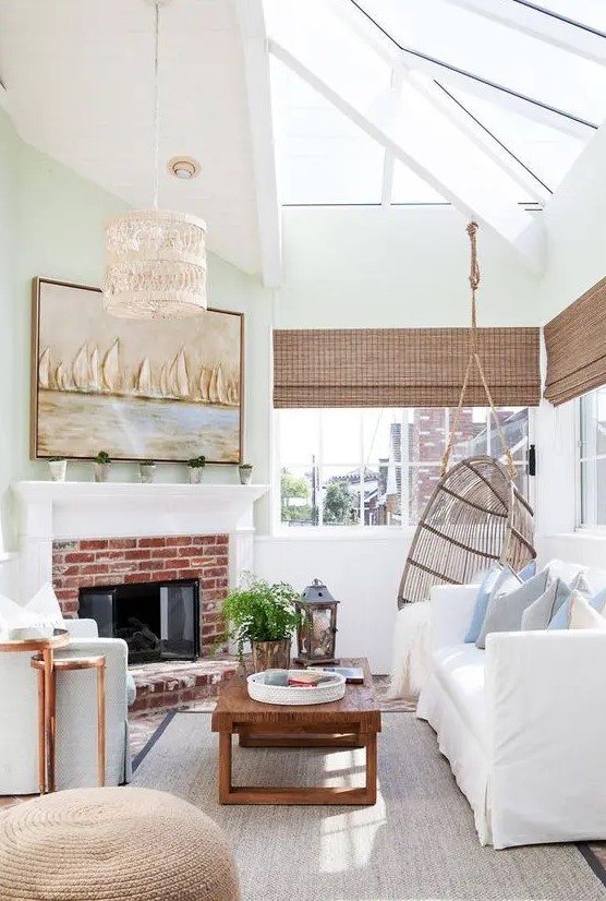 an elegant coastal sunroom with white and light blue furniture, woven shades, a brick fireplace, a woven lamp and a coastal artwork