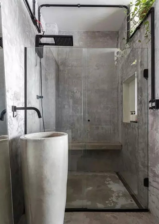 a minimalist industrial bathroom fully clad with concrete, a free standing sink, black pipes and potted greenery