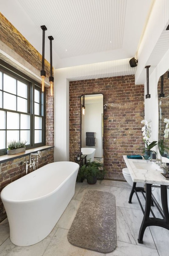 a modern industrial bathroom with brick walls, a stone sink on a stand, a free-standing tub, exposed pipes and bulbs