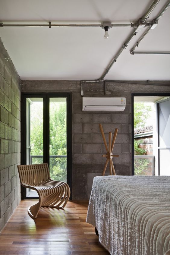 an industrial bedroom with brick walls, exposed pipes, a wooden chair and a rack plus a large bed with neutral bedding