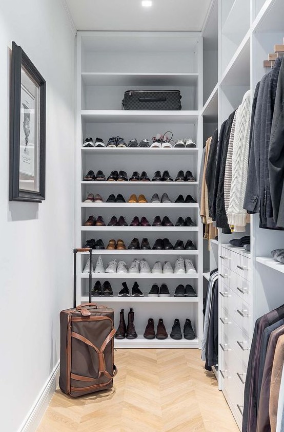 a small and stylish manly closet in white, with lots of open shoe shelves, holders with hangers, built-in drawers and lights