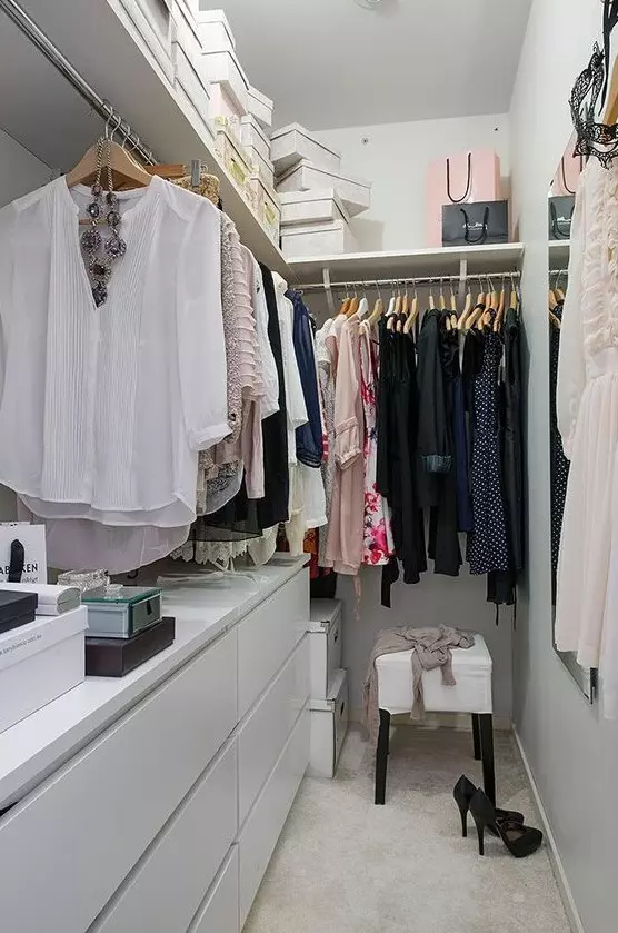 a small contemporary closet in white, with a large open shelf with boxes, dressers, holders with hangers and a stool