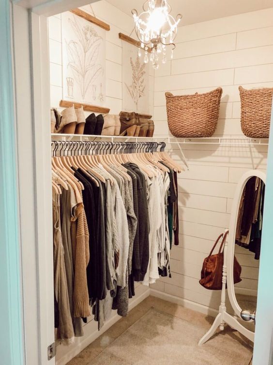 a small rustic closet with planked walls, a rack with clothes and a shoe shelf, a mirror and some botanical artwork