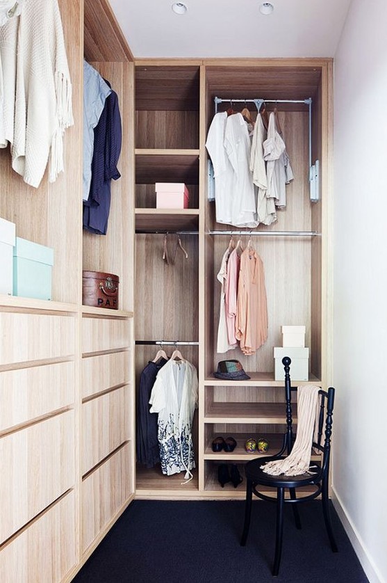 a small stylish closet done in light stained wood, with holders for clothes hangers, some open shelves, built in drawers and a black chair