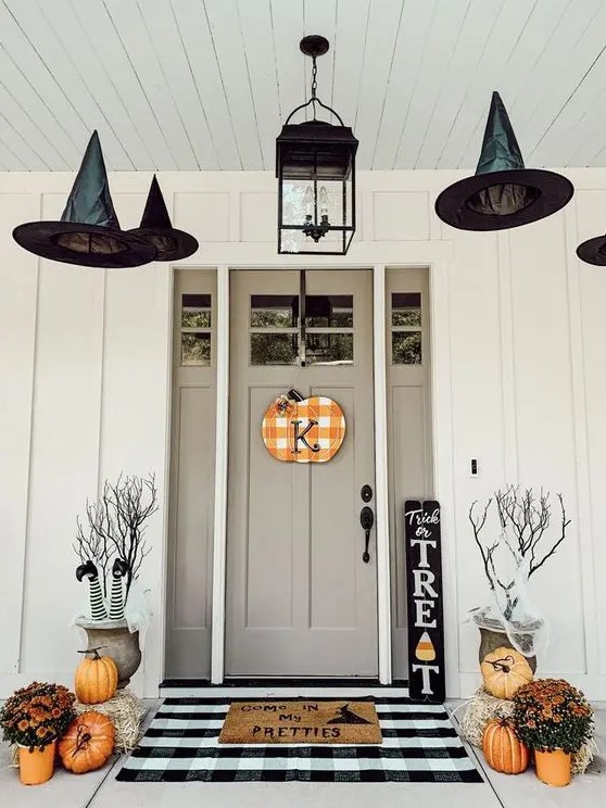 a Halloween porch decorated with witches' hats and legs, branches, pumpkins, hay, blooms and a plaid sign on the door