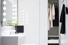 41 a white minimalist closet with a built-in wardrobe with sliding doors and drawers, a vanity with lights and a pouf for sitting