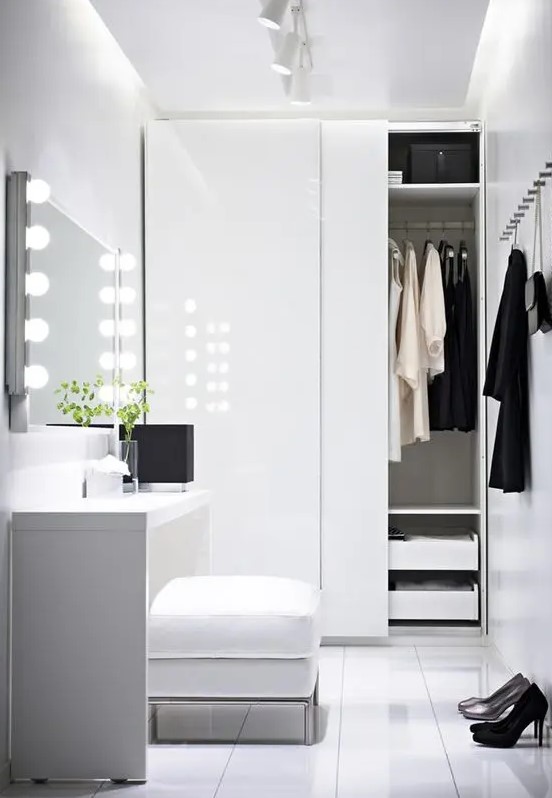 a white minimalist closet with a built in wardrobe with sliding doors and drawers, a vanity with lights and a pouf for sitting