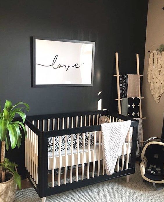 a boho nursery with a black accent wall, a black and white crib, a ladder for towels, a potted plant and a woven hanging