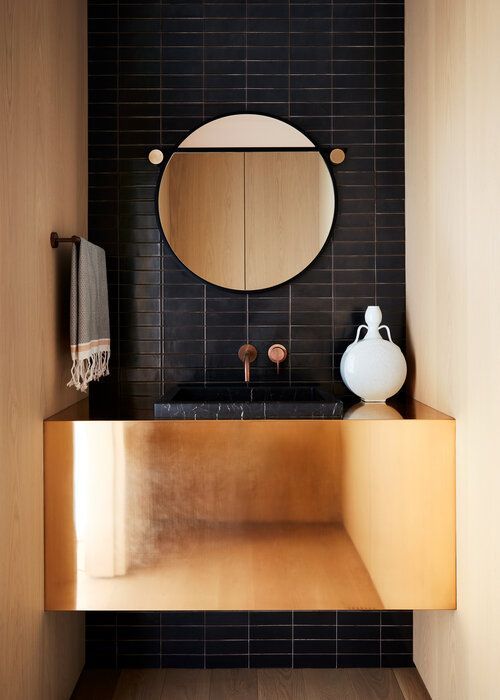 a bold bathing nook with plywood and a black skinny tile wall, a polished gold vanity, a round mirror and gold fixtures