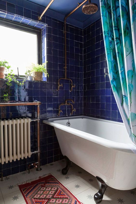 a bold bathroom with electric blue tiles, a clawfoot bathtub, a modern radiator, a bold boho rug and some potted plants