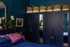 a bold eclectic bedroom with navy walls, navy wardrobes, baskets, a bed with electric blue bedding, a mirror and gold lamps