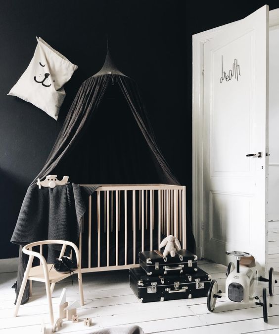 a bold monochromatic nursery with black walls, a crib with black bedding and a canopy, black suitcases, a stool and some toys