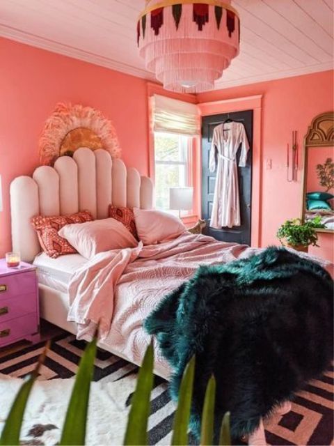 a bright eclectic bedroom with coral walls, a blush upholstered bed with pink bedding, a fringe chandelier, hot pink nightstands, potted plants