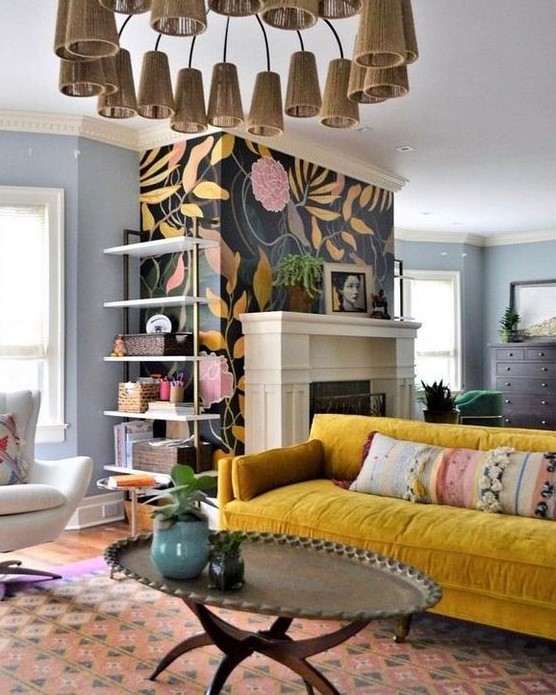 a bright eclectic living space with a unique chandelier, a mustard velvet sofa, a boho rug, a floral fireplace wall