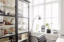 a catchy Scandinavian home office with a large glass storage unit, a white desk, a black chair, potted plants and artwork