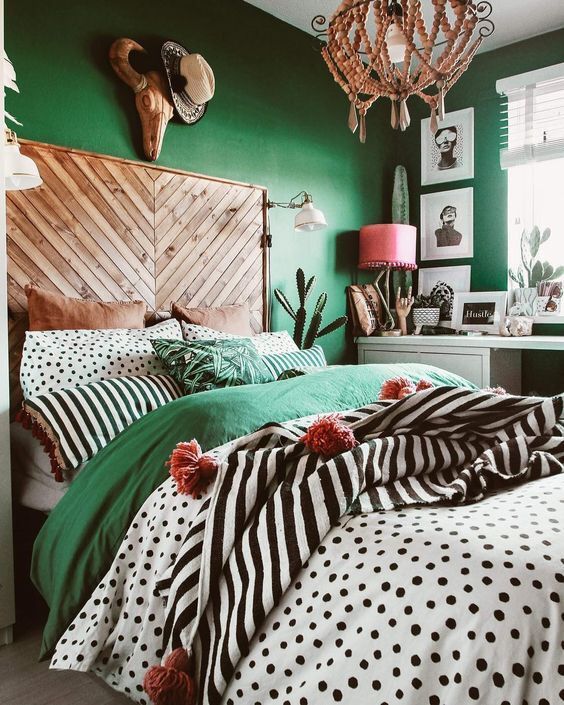 a catchy eclectic bedroom with emerald walls, a bed with a wooden headboard, colorful and printed bedding, a beaded chandelier, a pink lamp and some cacti