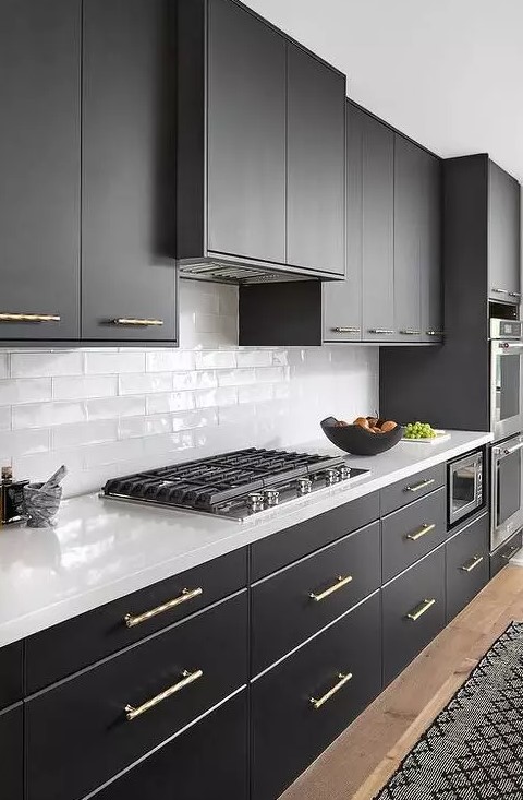 a chic black and white kitchen with sleek and matte black cabinets, white countertops, white skinny tiles on the backsplash and gold fixtures