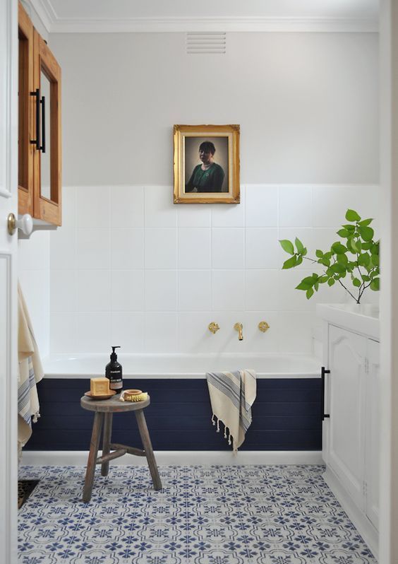 a chic eclectic bathroom with white square tiles and bold blue and white printed ones, a tub clad with navy tiles, a white cabinet and artwork