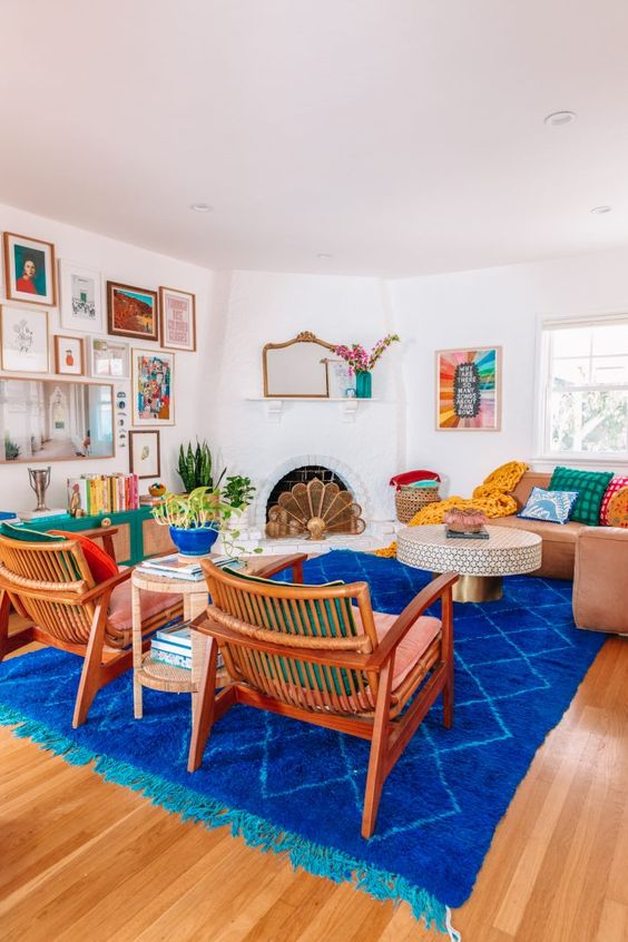 a colorful eclectic living room with a fireplace, a coffee table, a couple of pink chairs, a super bold rug and an emerald credenza