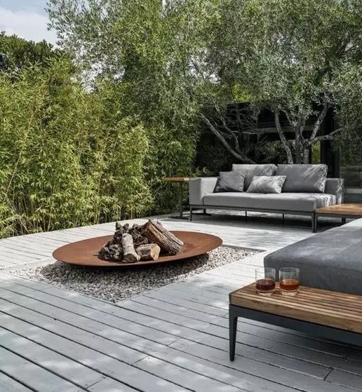 a contemporary outdoor space with grey sofas and wooden side tables, a greyish deck and a fire bowl in its center