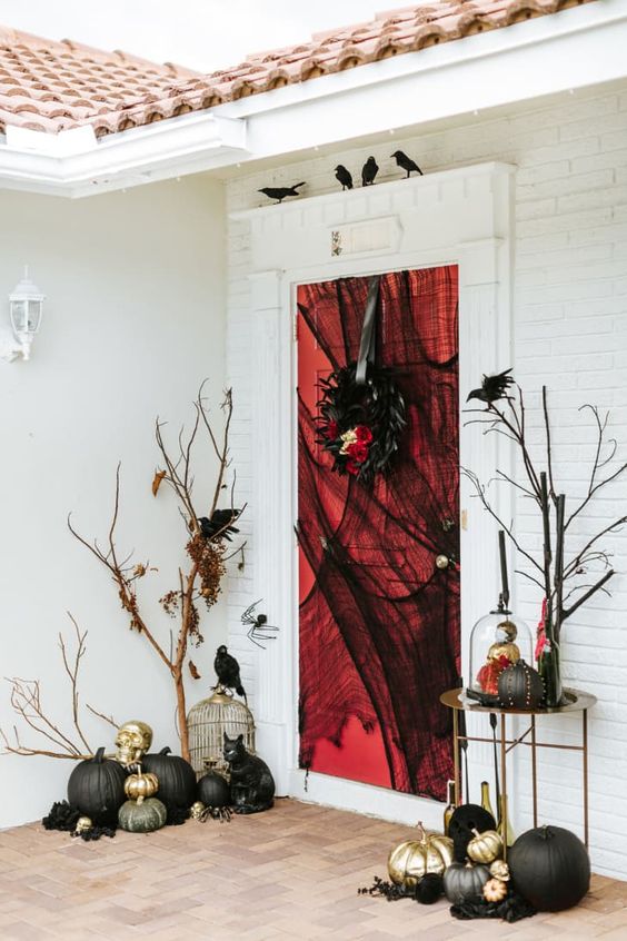 a cool black, gold and red Halloween porch with black cheesecloth, black and gold pumpkins, a cage, faux birds and hats and a wreath