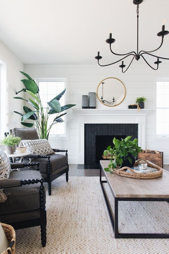 a cool farmhouse living room with a fireplace with a black surround, black vintage chairs, a coffee table, potted greenery and a black chandelier