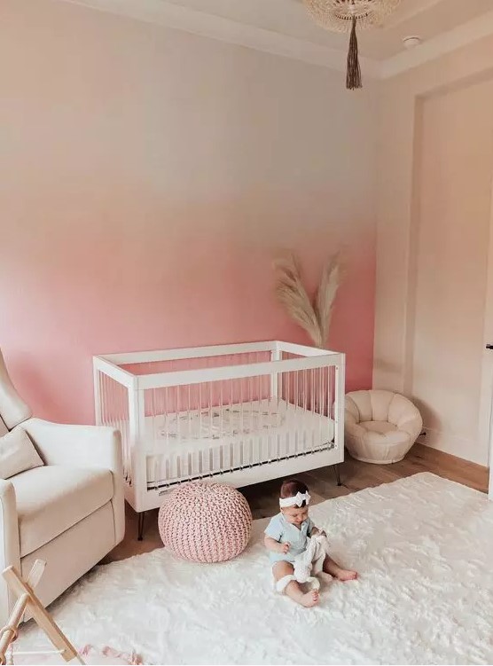 a cozy nursery with an ombre pink accent wall, creamy furniture, creamy textiles and a pink chunky knit pouf is wow