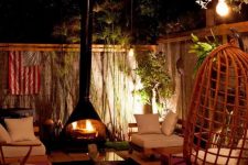 a cozy outdoor living room with a black Malm fireplace, neutral seating furniture, a rug, an egg pendant chair and pendant bulbs