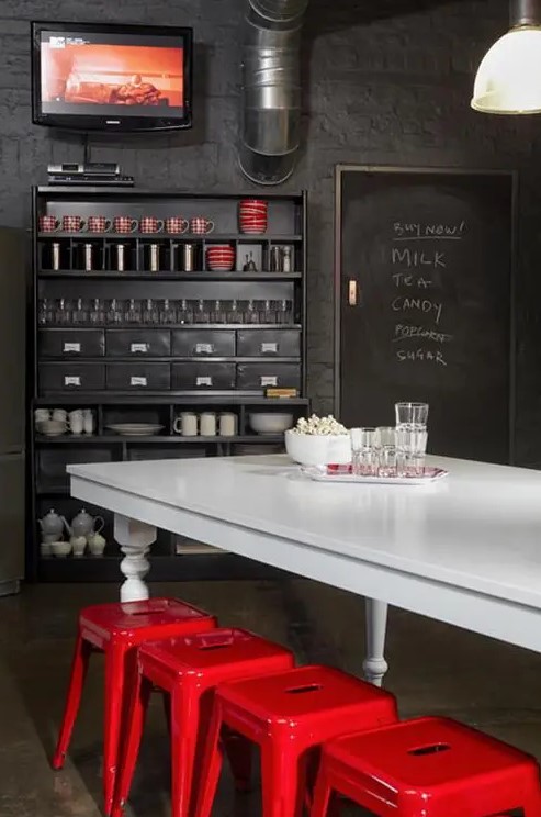 a dark and moody industrial dining space with black walls, a black storage unit, exposed pipes, a white vintage table and hot red metal chairs