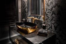 a glam and chic bathroom with black wallpaper, a black marble vanity, a black and gold sink, a mirror in a gold frame