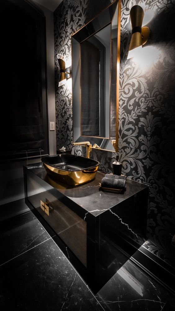 a glam and chic bathroom with black wallpaper, a black marble vanity, a black and gold sink, a mirror in a gold frame