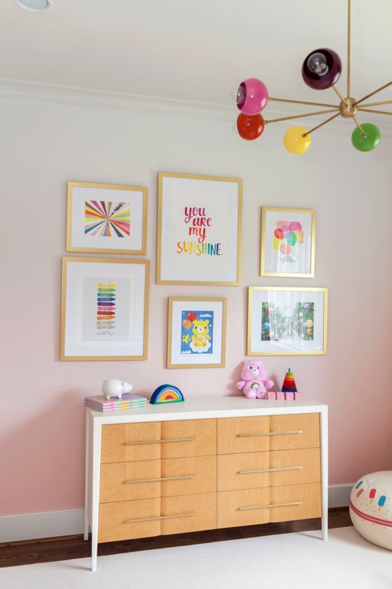 a kid's space with an ombre pink accent wall, a stained dresser, a colorful gallery wall and bright toys plus a colorful chandelier
