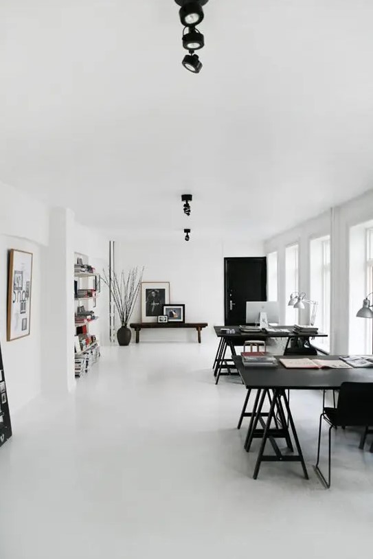 a large black and white Scandinavian home office with two large black desks and black chairs, a wooden bench, bookshelves and silver table lamps plus lots of natural light