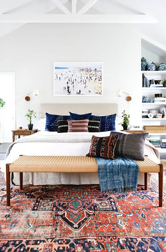 a light-filled eclectic bedroom with an upholstered bed, a woven bench, built-in shelves, wooden nightstands and potted greenery