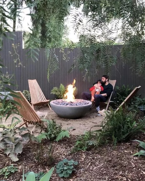 a lovely fire pit with a large stone ethanol fire bowl with pebbles and wooden chairs and greenery around is amazing