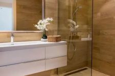 a minimalist bathroom fully clad with wood, with a glass-enclosed shower space, a floating white vanity and a mirror